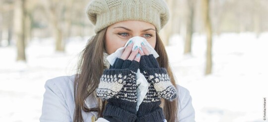 Frau im Winter mit schneebedeckter Landschaft hält sich ein Taschentuch an die Nase 