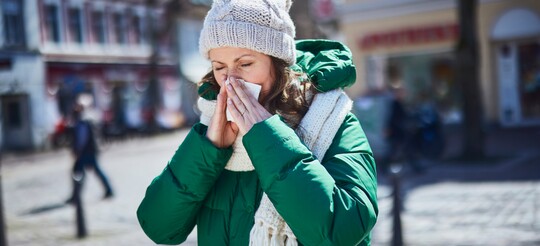 Frau im Winter mit laufender Nase, verursacht durch Kälte und trockene Luft. Eine Frau in einer dicken grünen Winterjacke und weißer Mütze steht auf einer winterlichen Straße und schnäuzt sich in ein Taschentuch. Im Hintergrund sind unscharfe Gebäude und Menschen zu sehen.