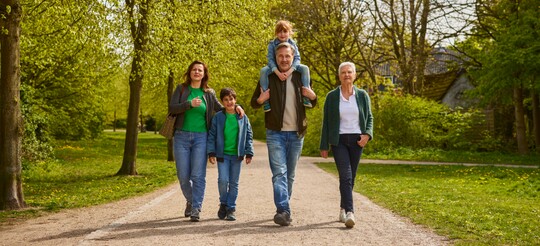 Gesunde Atemwege für alle Generationen: Familie genießt Spaziergang im Grünen Eine multigenerationale Familie aus fünf Personen spaziert lächelnd auf einem Waldweg. Ein älterer Mann trägt ein Kind auf den Schultern. Umgeben von grünen Bäumen symbolisiert die Szene Gesundheit und Wohlbefinden in der Natur.