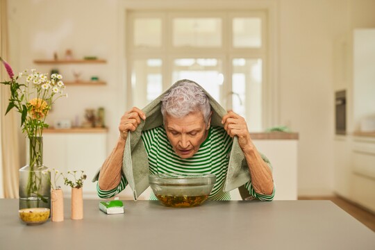 Inhalation zur Linderung einer trockenen und verstopften Nase. Ältere Frau mit grauen Haaren inhaliert unter einem Handtuch über einer Schüssel mit heißem Wasser und Kräutern, umgeben von Blumen und Pflegeprodukten.