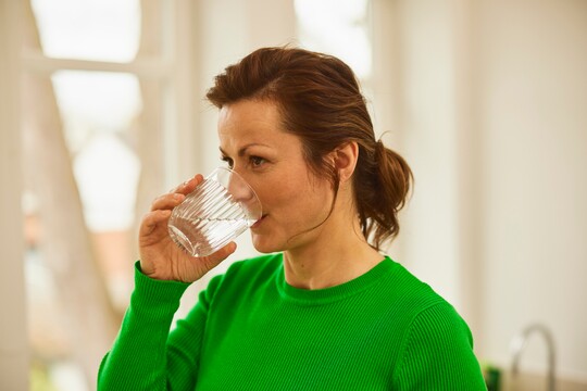 Frau trinkt Wasser zur Unterstützung der Feuchtigkeit bei trockener Nase. Frau mittleren Alters mit braunen Haaren trinkt ein Glas Wasser in einem hellen Raum, um sich zu erfrischen.