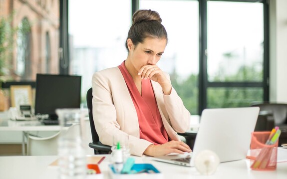 Junge Frau im Büro am Laptop, anscheinend mit einer Erkältung, hält sich die Hand an die Nase. im Vordergrund ist unscharf geloSitin zu sehen