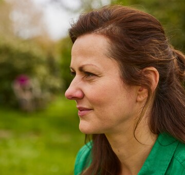 Nahaufnahme einer Frau im Profil, die im Freien in einem Garten sitzt. Sie trägt eine grüne Jacke und hat braune Haare. Ihr Gesichtsausdruck wirkt nachdenklich, möglicherweise aufgrund von Beschwerden durch eine trockene oder wunde Nase. Im Hintergrund sind unscharfe Bäume und Wiesen zu erkennen.