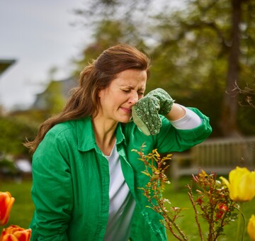 Eine Frau in einem grünen Hemd arbeitet im Garten und reibt sich die Nase mit einem Handschuh, möglicherweise aufgrund von Pollenallergie. Im Vordergrund sind Blumen zu sehen, und der Hintergrund zeigt Bäume und eine Bank im Garten.