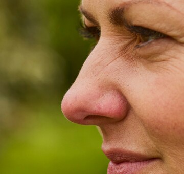 Nahaufnahme einer Frau im Profil, die im Freien in einem Garten sitzt. Sie trägt eine grüne Jacke und hat braune Haare. Ihr Gesichtsausdruck wirkt nachdenklich, möglicherweise aufgrund von Beschwerden durch eine trockene oder wunde Nase. Im Hintergrund sind unscharfe Bäume und Wiesen zu erkennen.